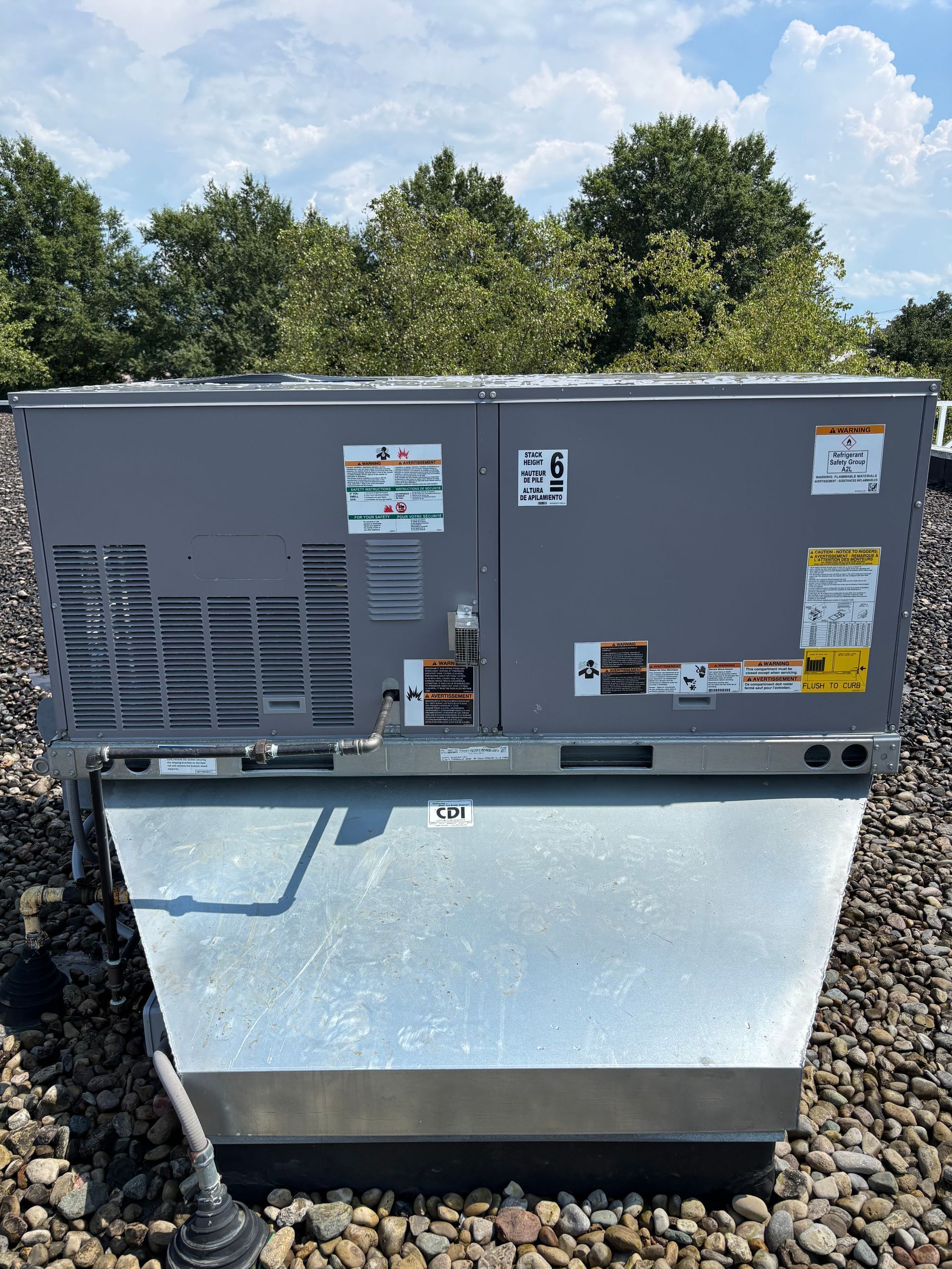 Rooftop HVAC unit on a gravel surface against a backdrop of trees and a partly cloudy sky.