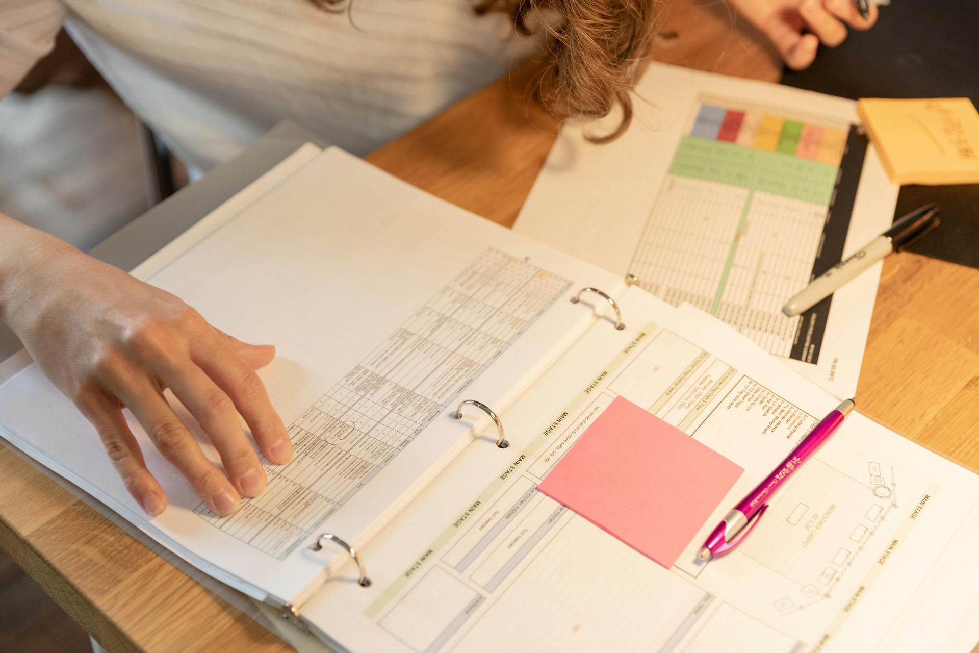 Une personne examine des documents dans un classeur, assise à un bureau, avec un post-it et un stylo.