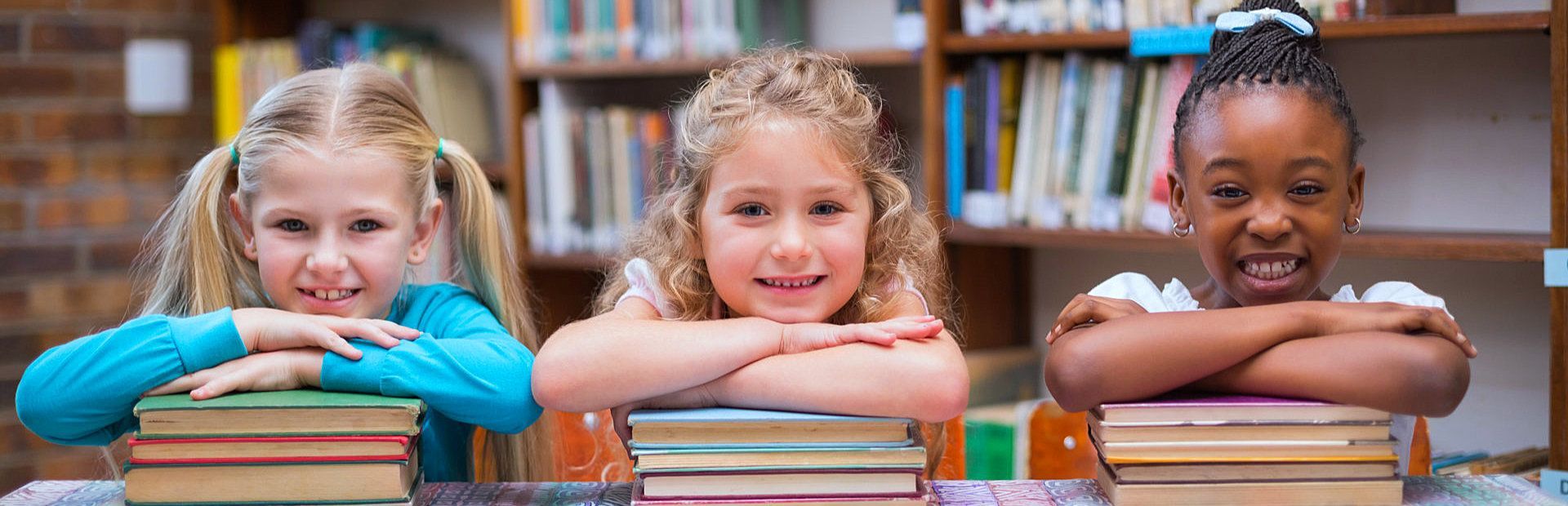 Three little girls are leaning on stacks of books in a library.