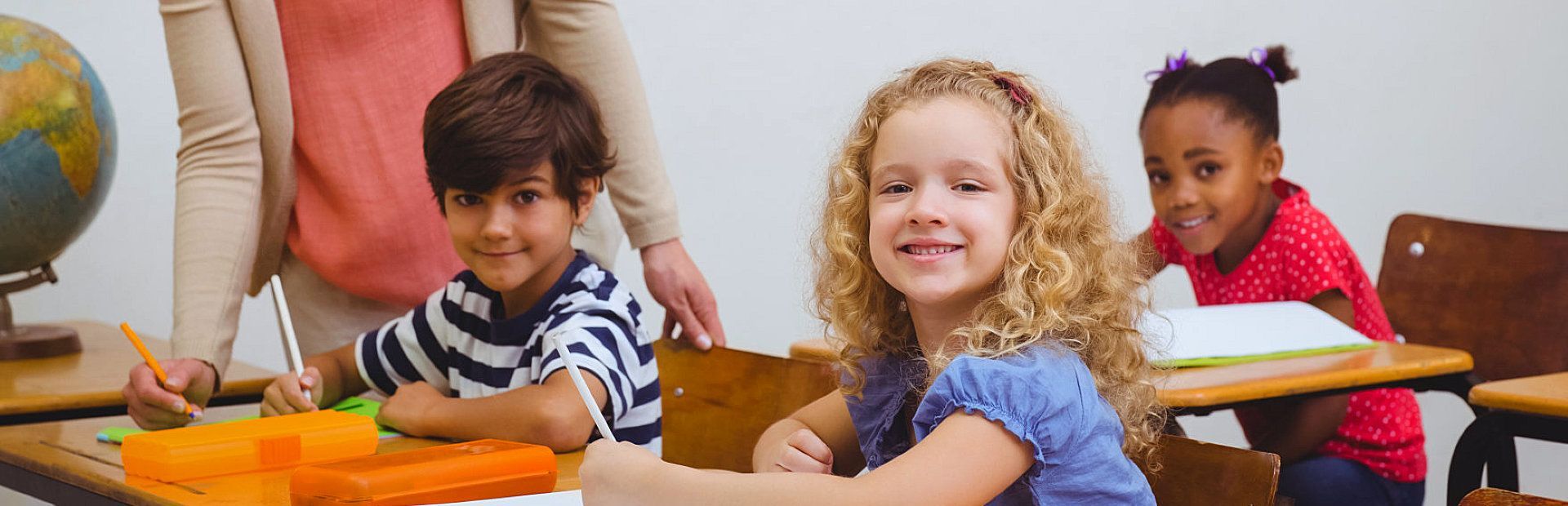 A group of children are sitting at desks in a classroom with their teacher.