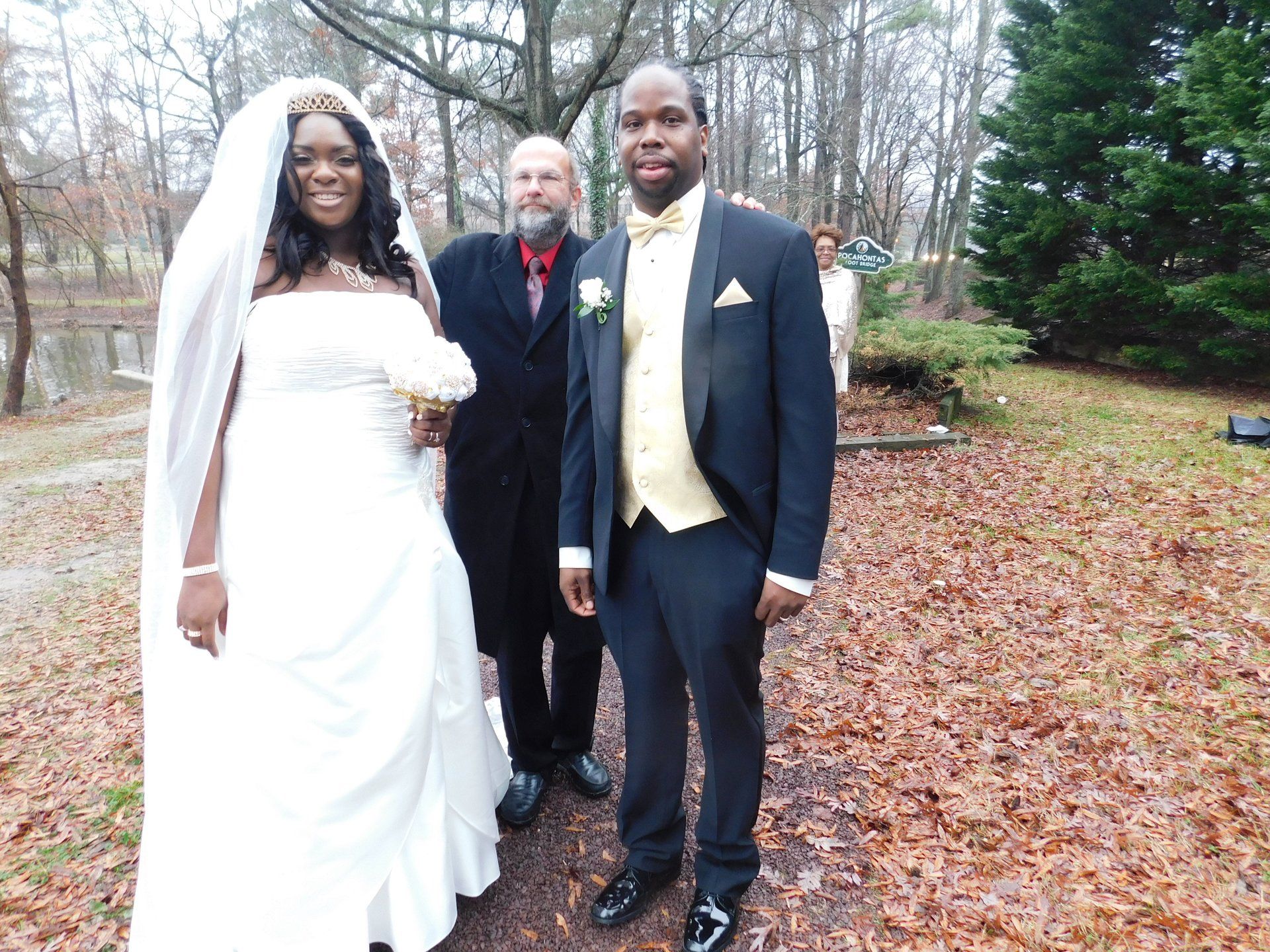 Bride and groom pose with officiant Rev. Byron Sichert in wooded area on wedding day. Bride wears white gown, groom a tuxedo.