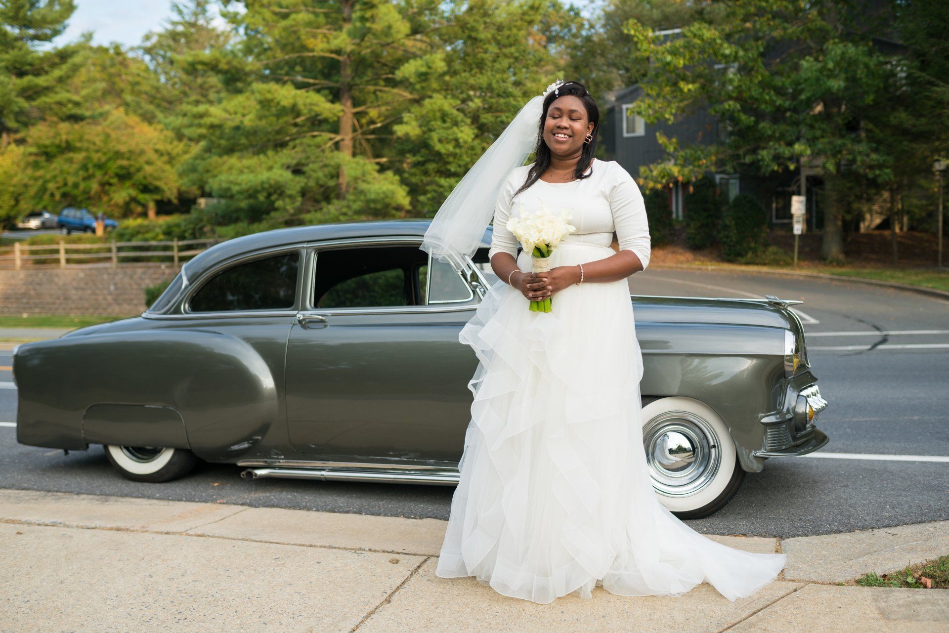 A woman in a wedding dress is standing in front of an old car.