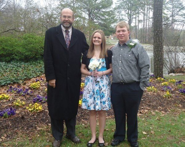 A man in a black coat stands next to a woman in a blue dress and a man in a grey shirt