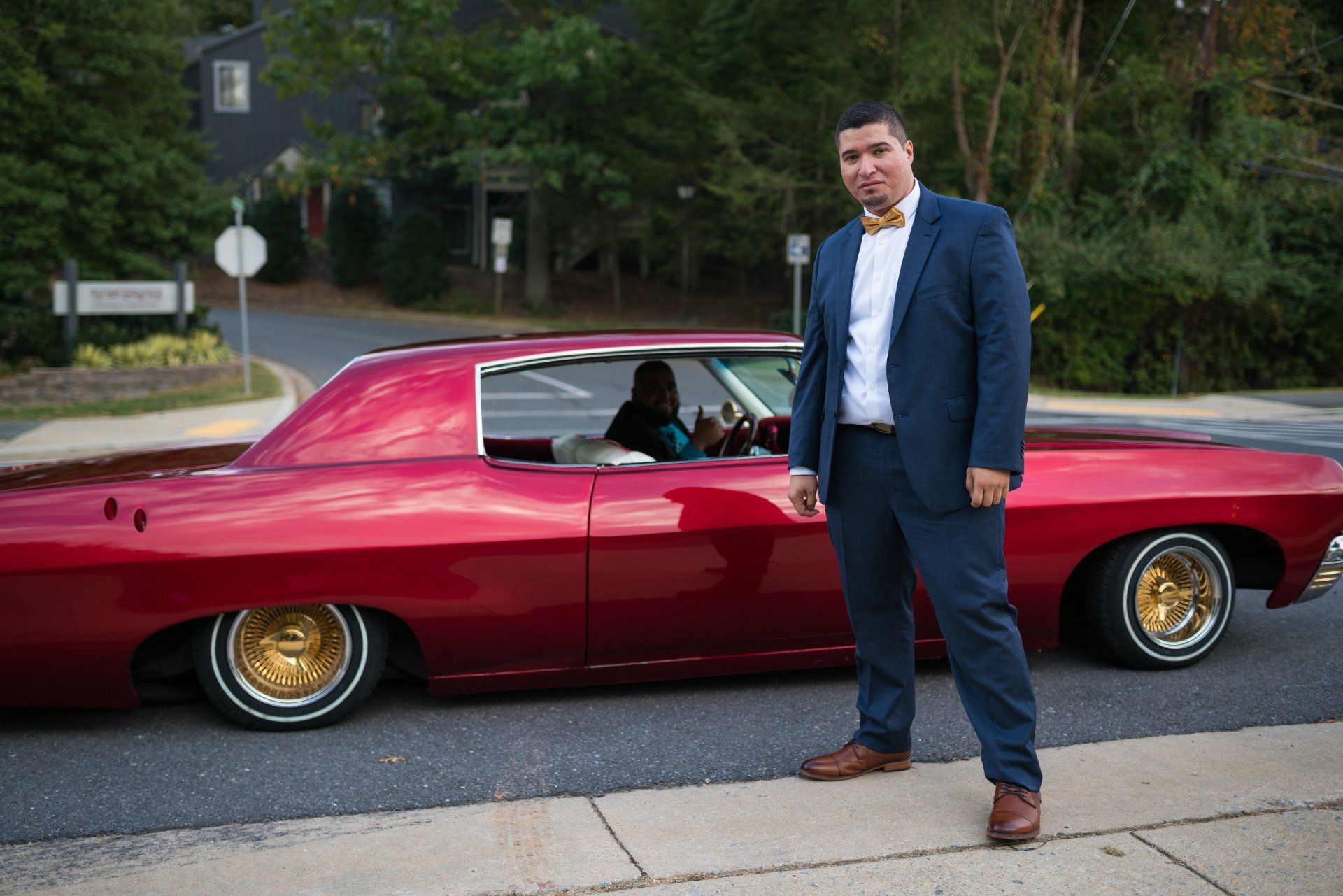 A man in a suit and bow tie is standing next to a red car.