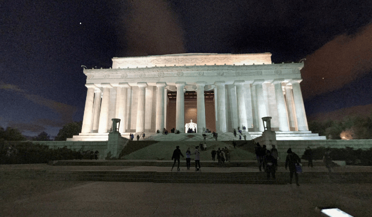 The lincoln memorial is lit up at night with people walking in front of it