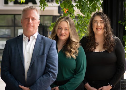Three team member of Martin IP Law Group standing outdoors, smiling warmly. They are under green leafy branches. The mood is professional and welcoming.