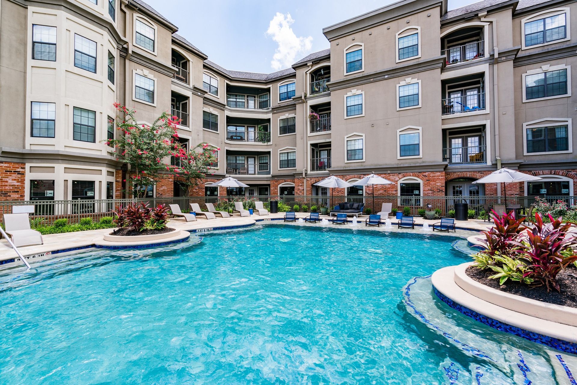 Apartment complex courtyard with pool. Beige buildings surround the pool with lounge chairs and umbrellas.