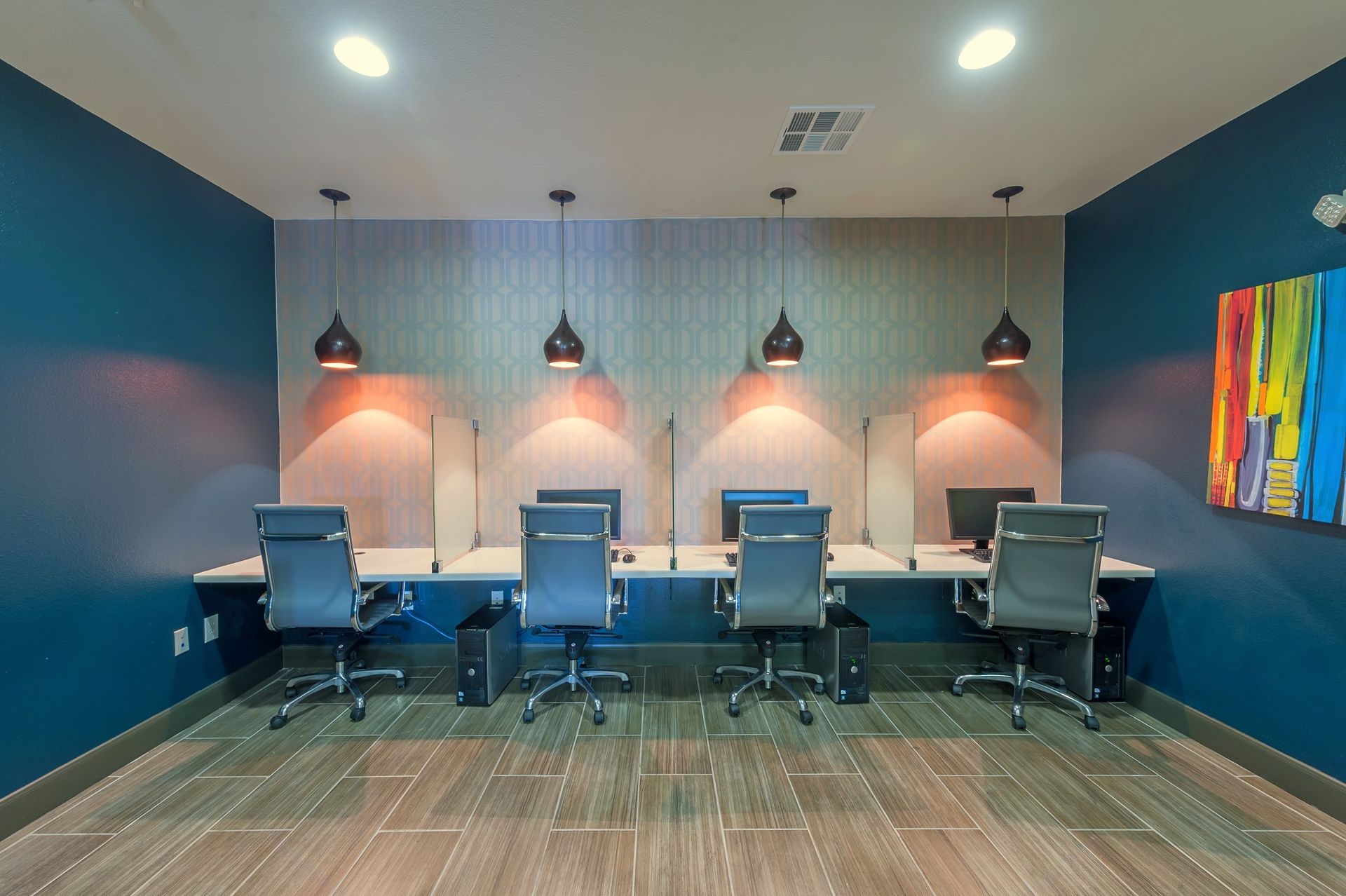Computer workstation in a teal-walled room with gray chairs, pendant lights, and colorful artwork.