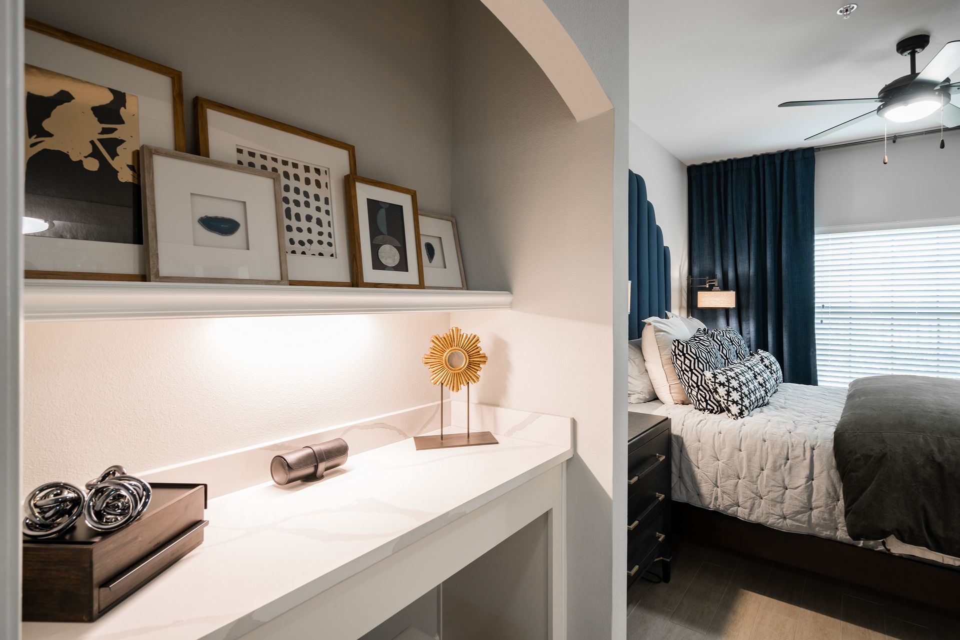 Nook with art above a white countertop and a bed with blue curtains in the background.