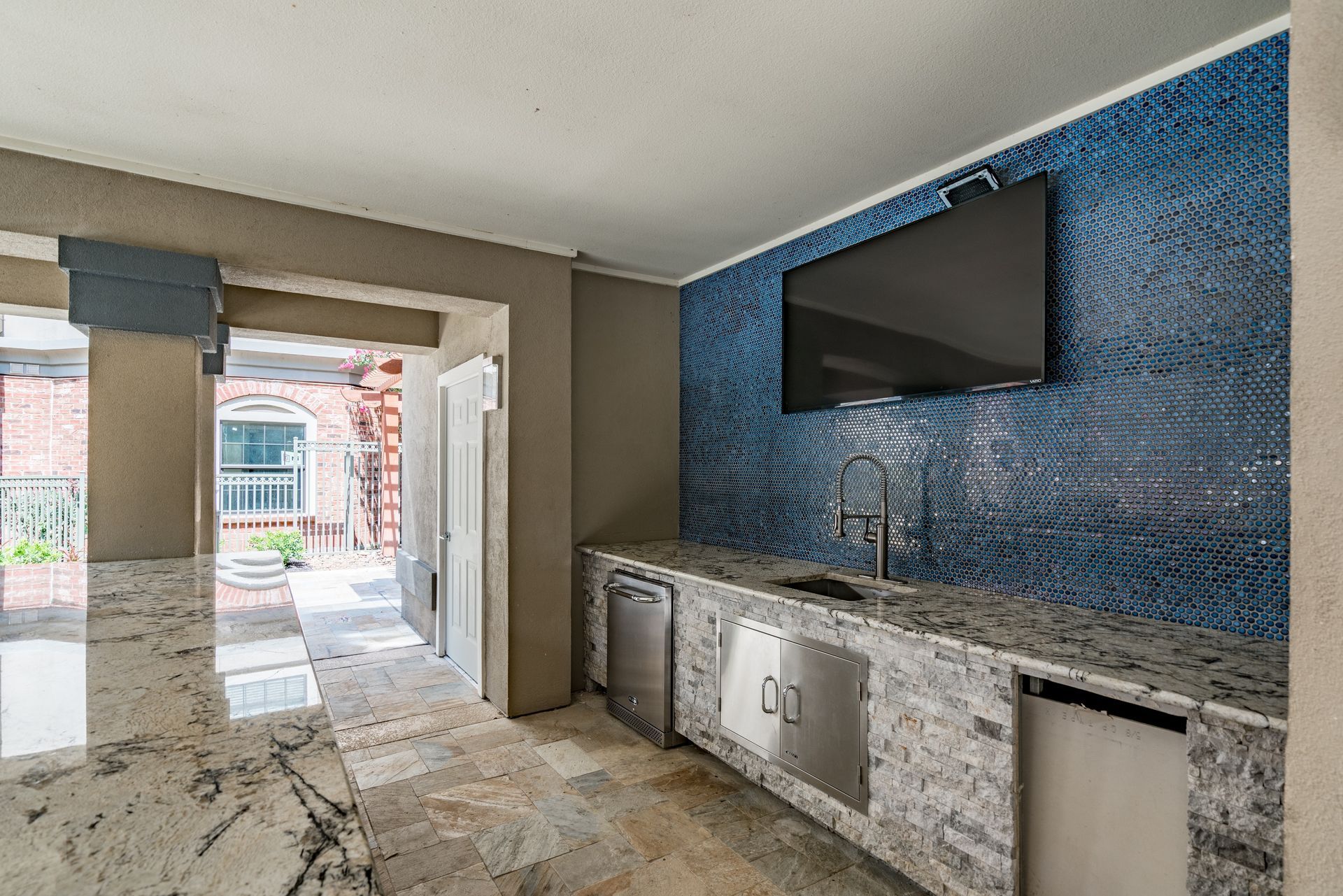 Outdoor kitchen with granite countertops, blue tiled backsplash, TV, and stainless steel appliances.