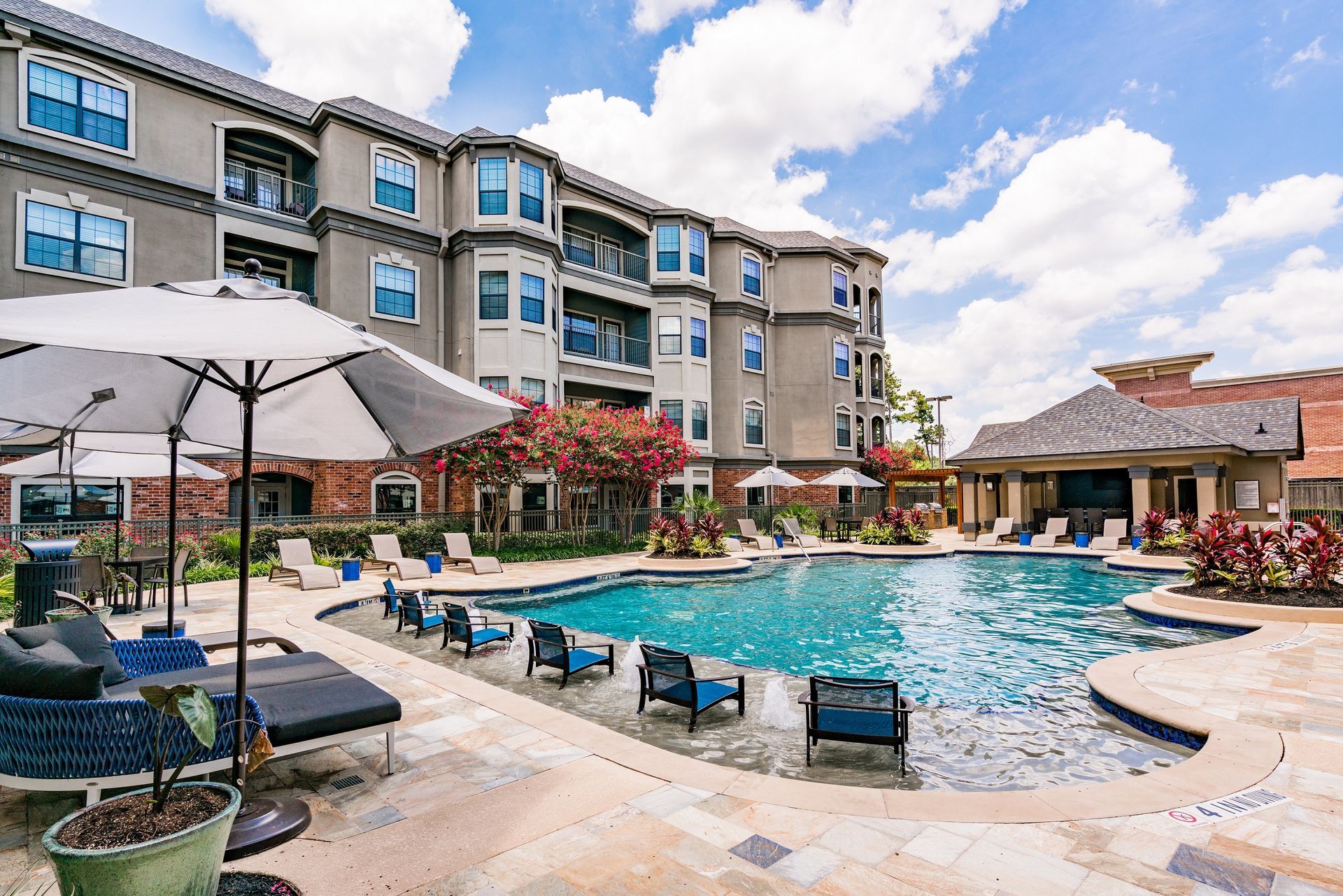 Apartment pool with lounge chairs, umbrellas, and a cabana under a blue sky.
