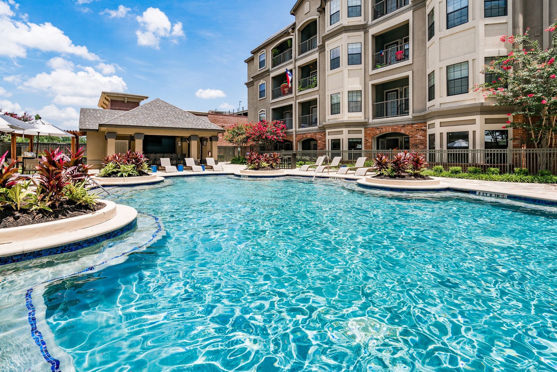 Swimming pool in front of an apartment building with a cabana. Blue water with lush landscaping.