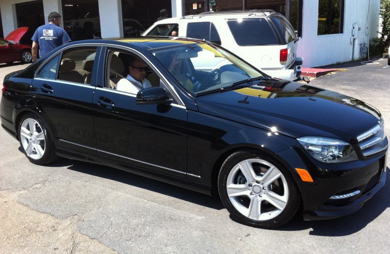 A black Mercedes-Benz sedan parked in a garage lot with a driver behind the wheel and a person standing behind the car. | Allstar Automotive