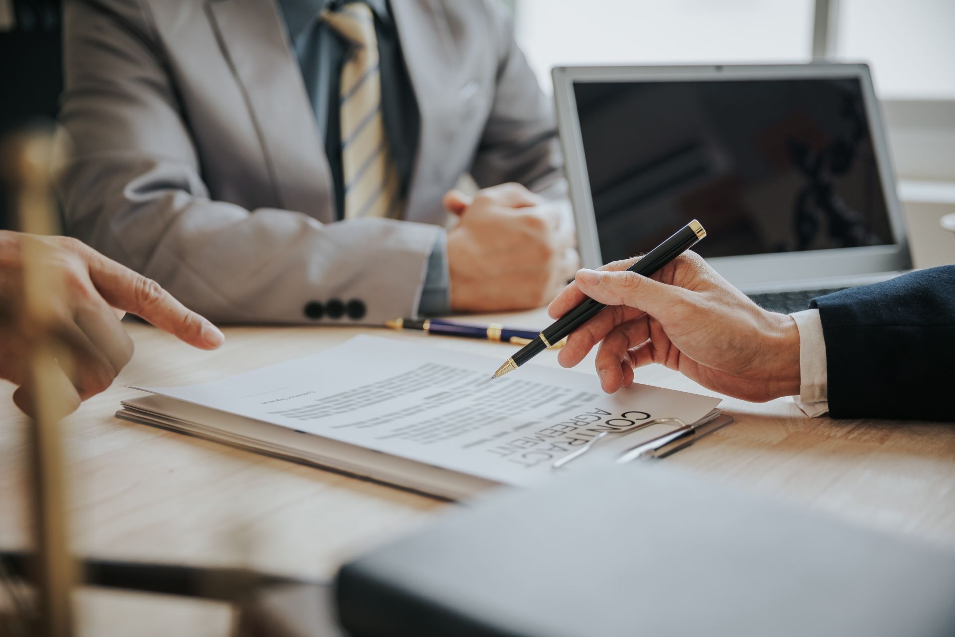 People in suits reviewing a document, one pointing with a finger, another holding a pen, laptop in background.