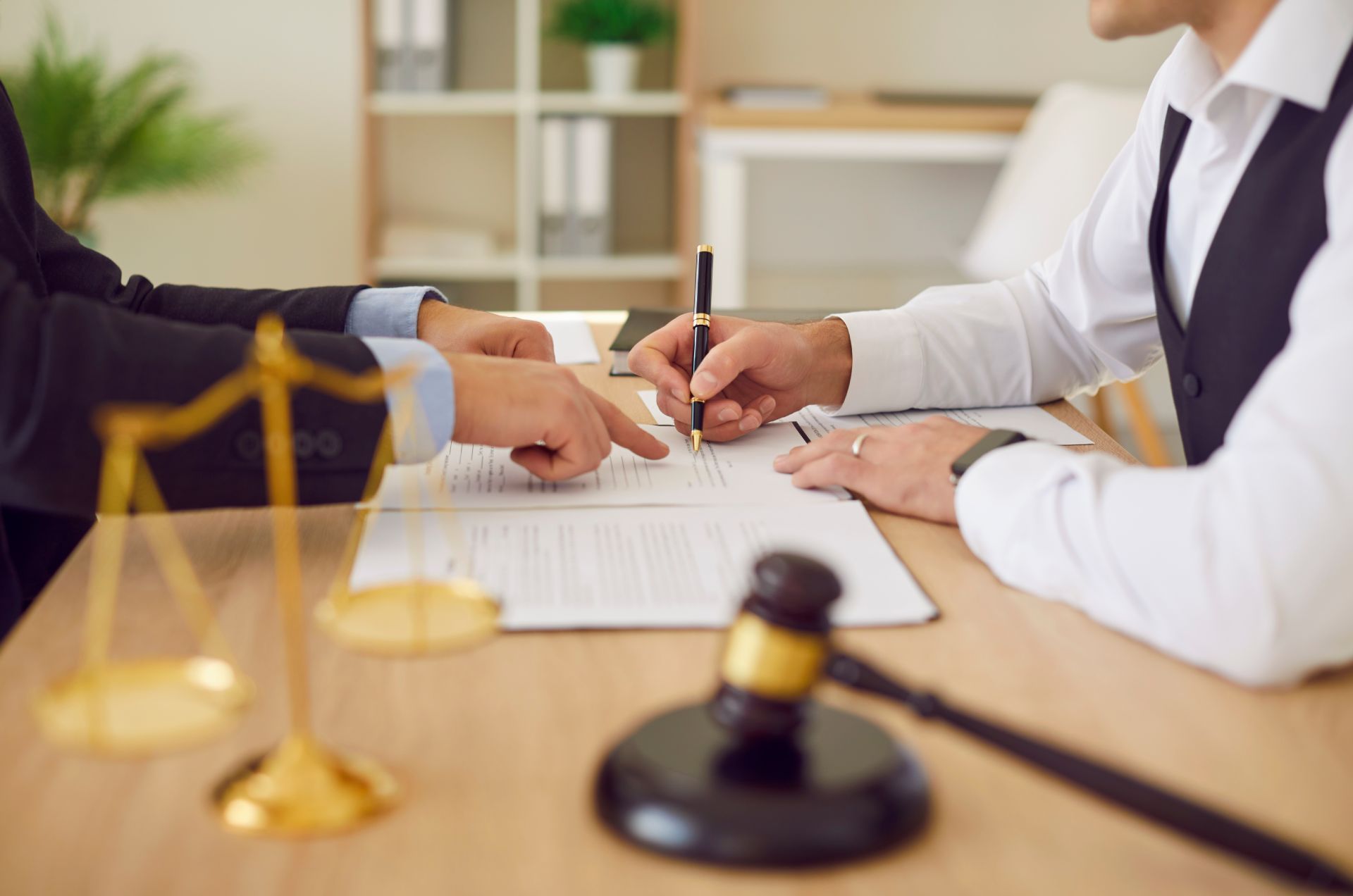 Lawyer pointing to a document as a client signs. Golden scale and gavel on table.
