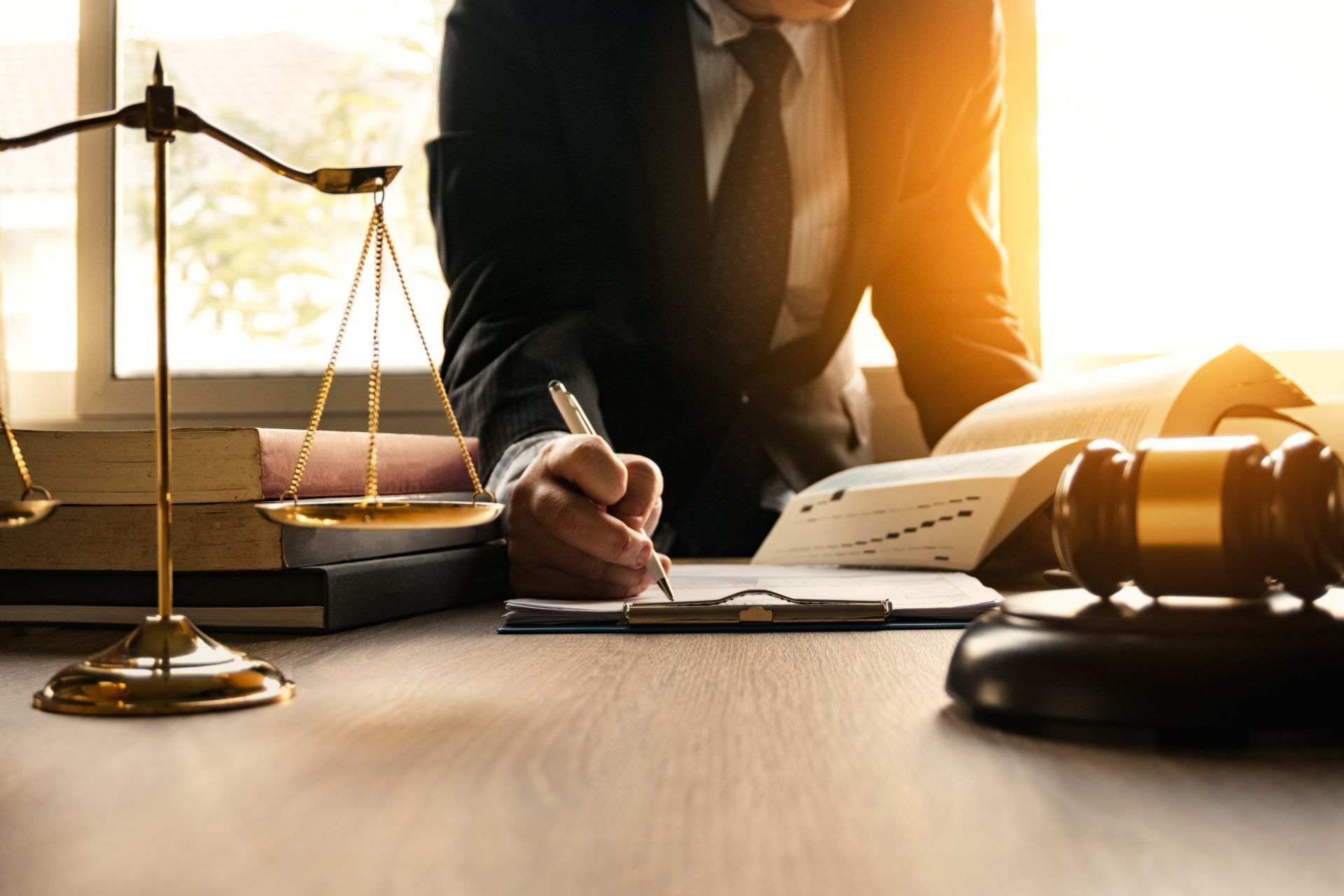 Lawyer in suit writing at a desk with scales, gavel, and books, lit by sunlight.