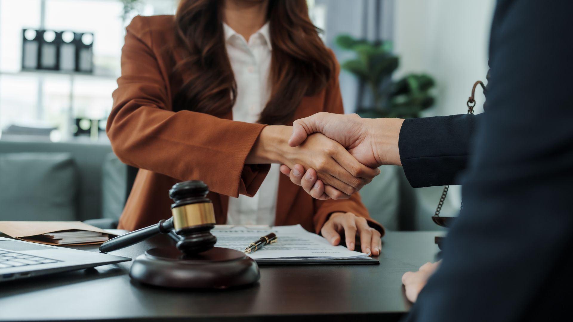 Woman in brown blazer shakes hands with a person in a suit, gavel and documents on the desk.
