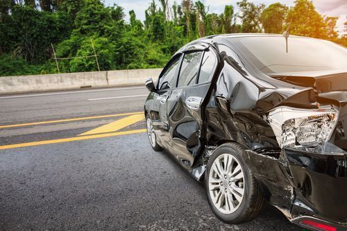 Damaged black car on roadside after collision; rear side severely dented; highway in background.