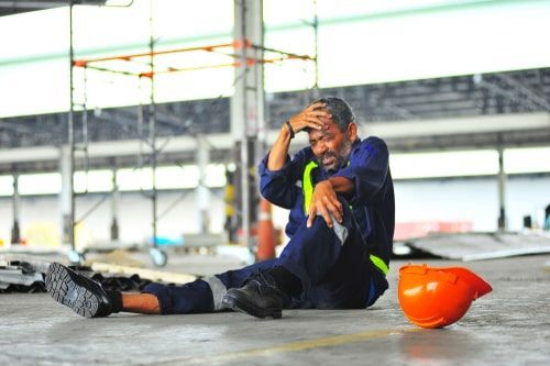 Worker in blue uniform sitting on the ground, holding his head; orange helmet nearby.