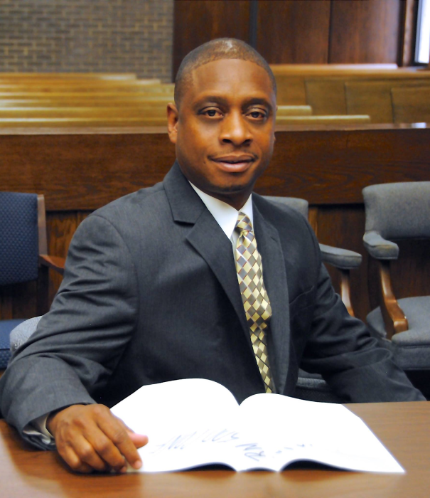 Man in suit seated at a table, holding open book in a courtroom setting.