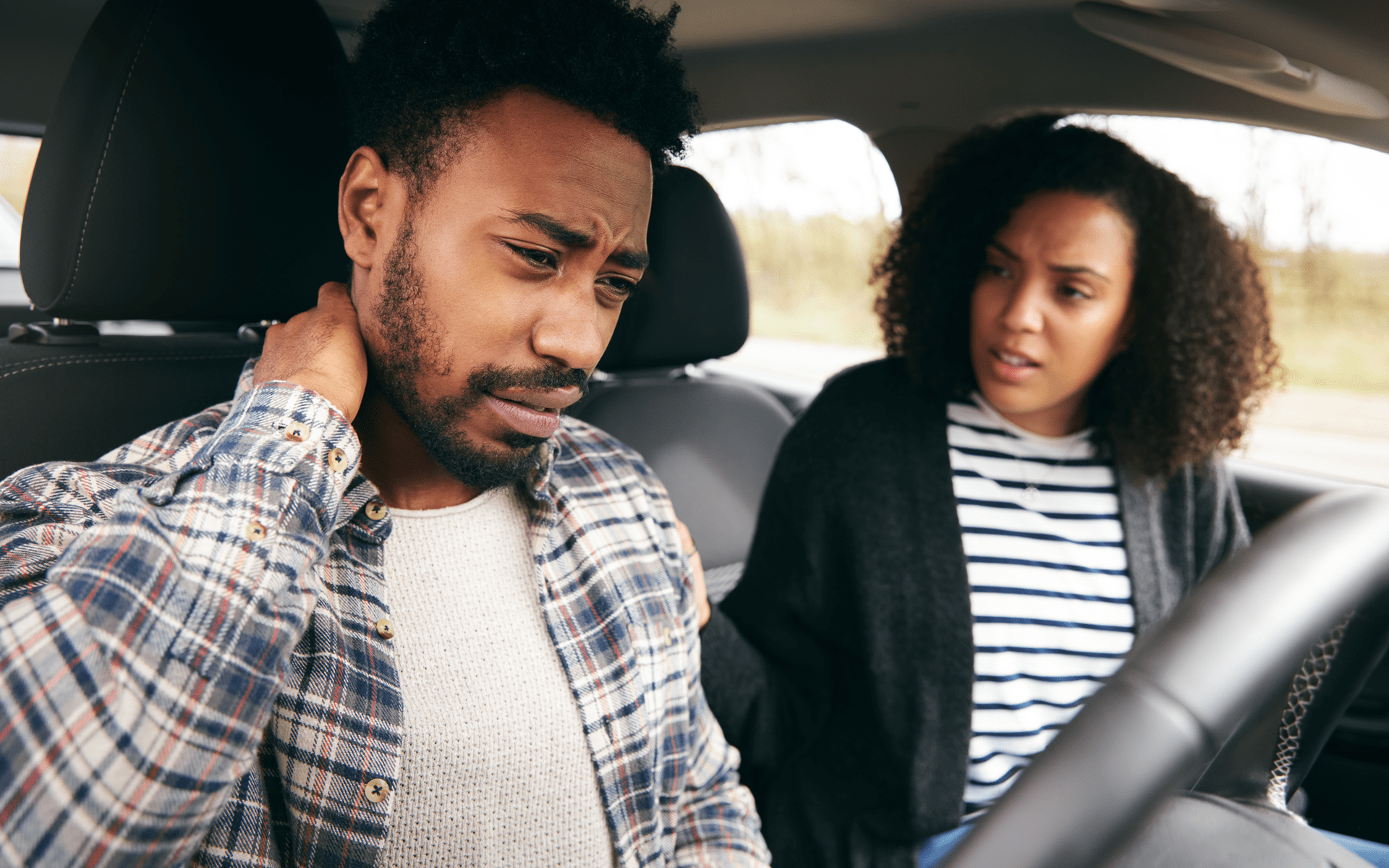 Man in a plaid shirt clutches neck, sitting in car. Woman looks concerned beside him.