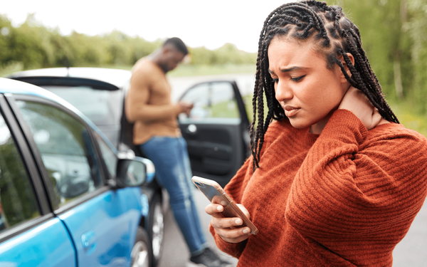 Woman holding her neck after a car accident, checking her phone. Man leans on a damaged car in the background.
