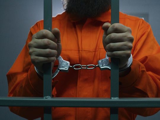 A man in an orange shirt and handcuffs is behind bars.
