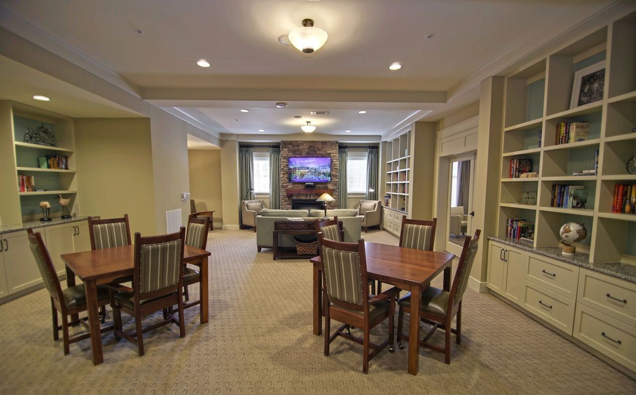 Interior of a community lounge with tables, chairs, built-in bookshelves, and a stone fireplace with a TV.