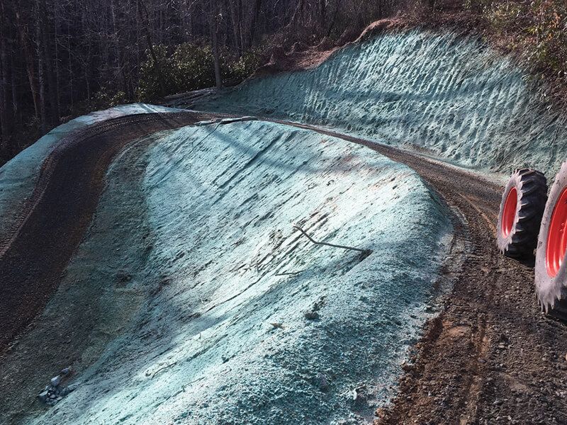 A tractor is driving down a dirt road next to a hill covered in grass.