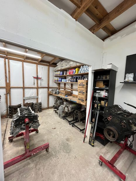An auto repair workshop with engine parts on stands, shelves of supplies, and a concrete floor.
