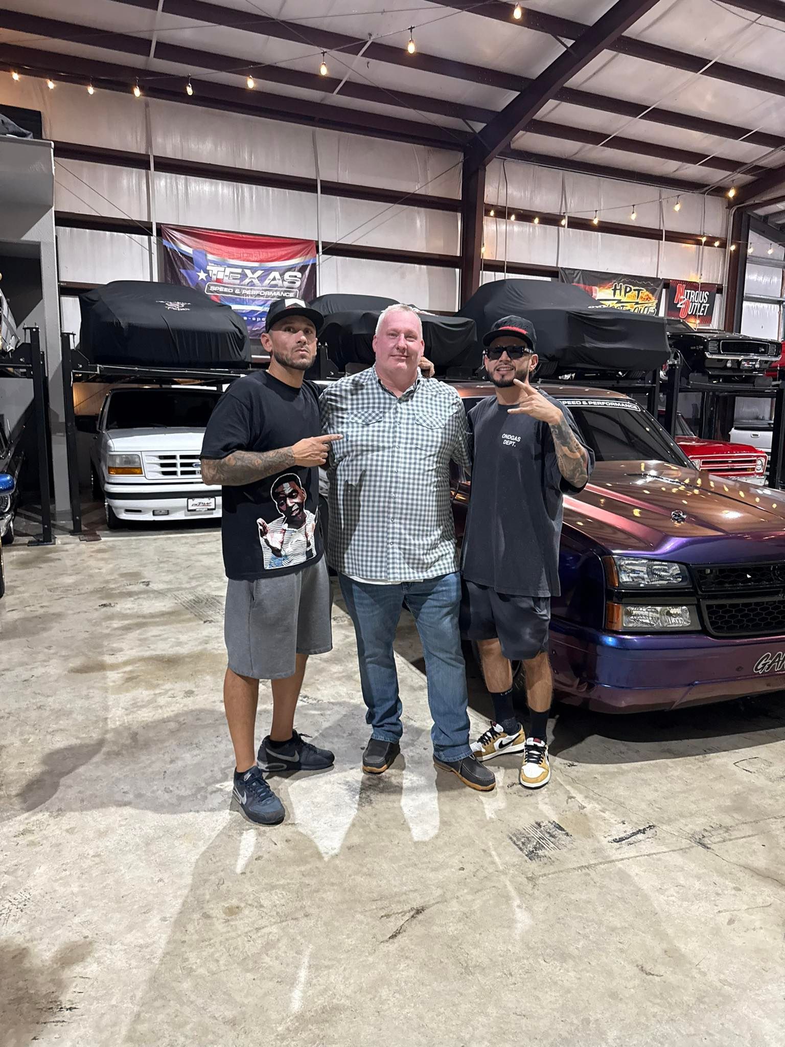 Three men pose in a garage with custom trucks. Two wear hats, with one having arm tattoos. performance parts and drag truck.