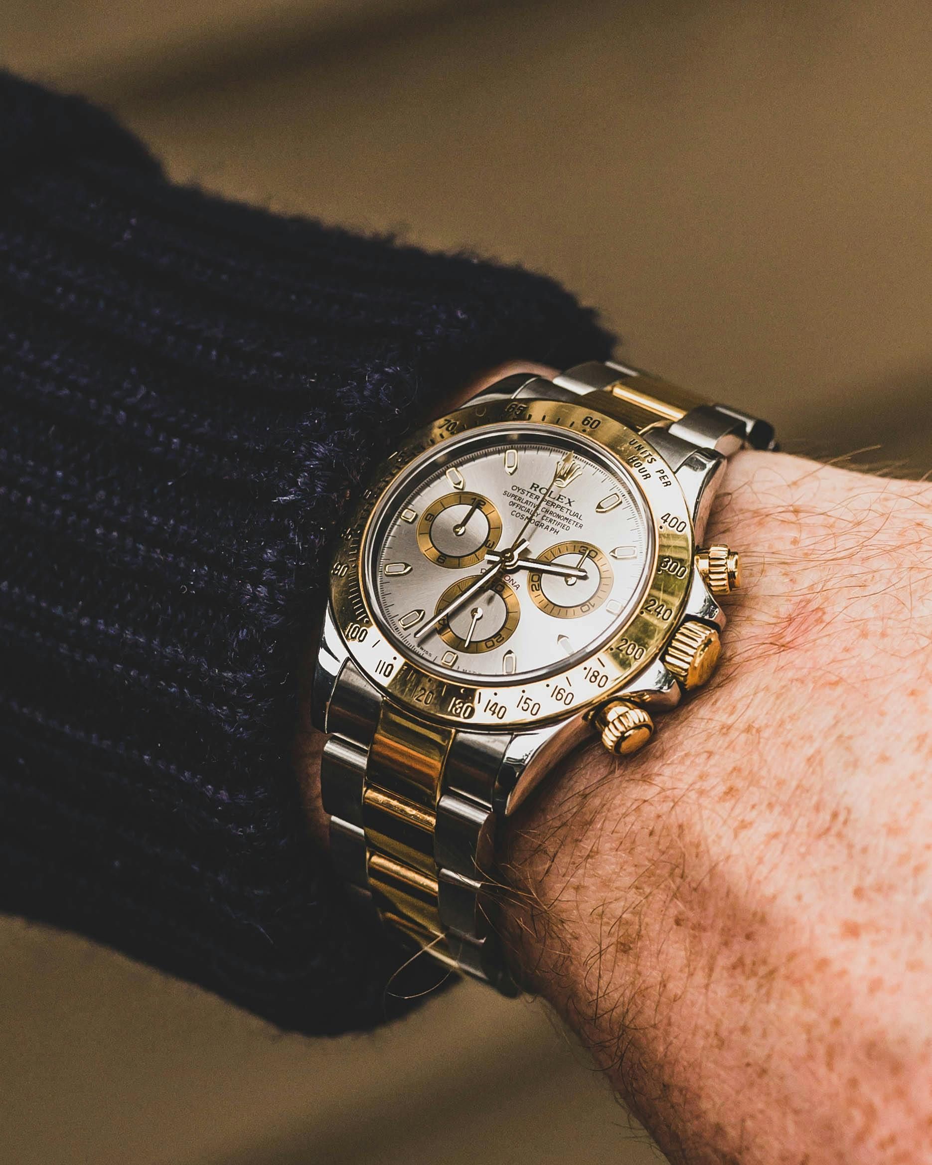 Wrist with two-tone Rolex Daytona watch, silver dial, gold bezel and bracelet, black sweater sleeve.
