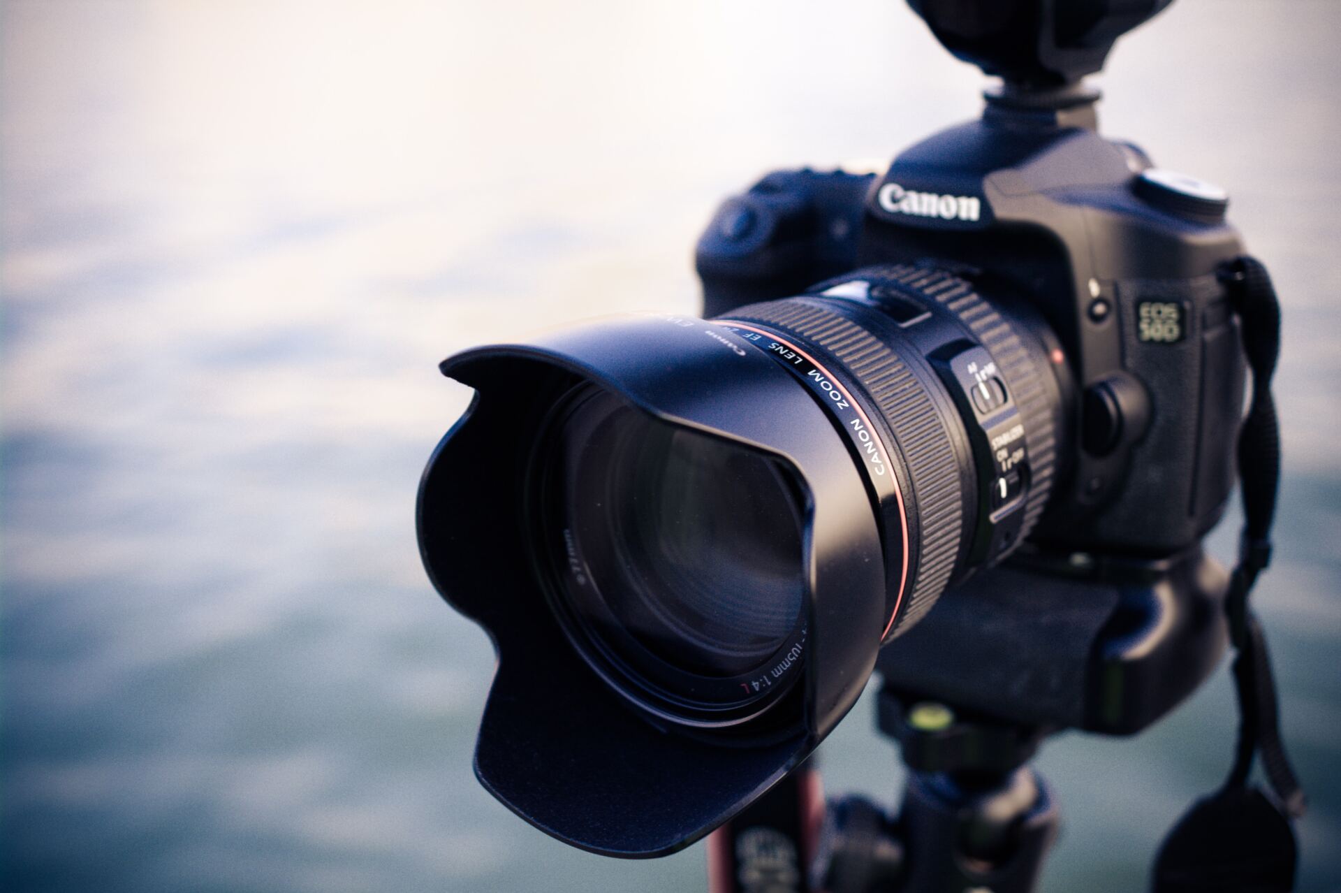 Canon DSLR camera with lens hood mounted on a tripod, blurred water background.