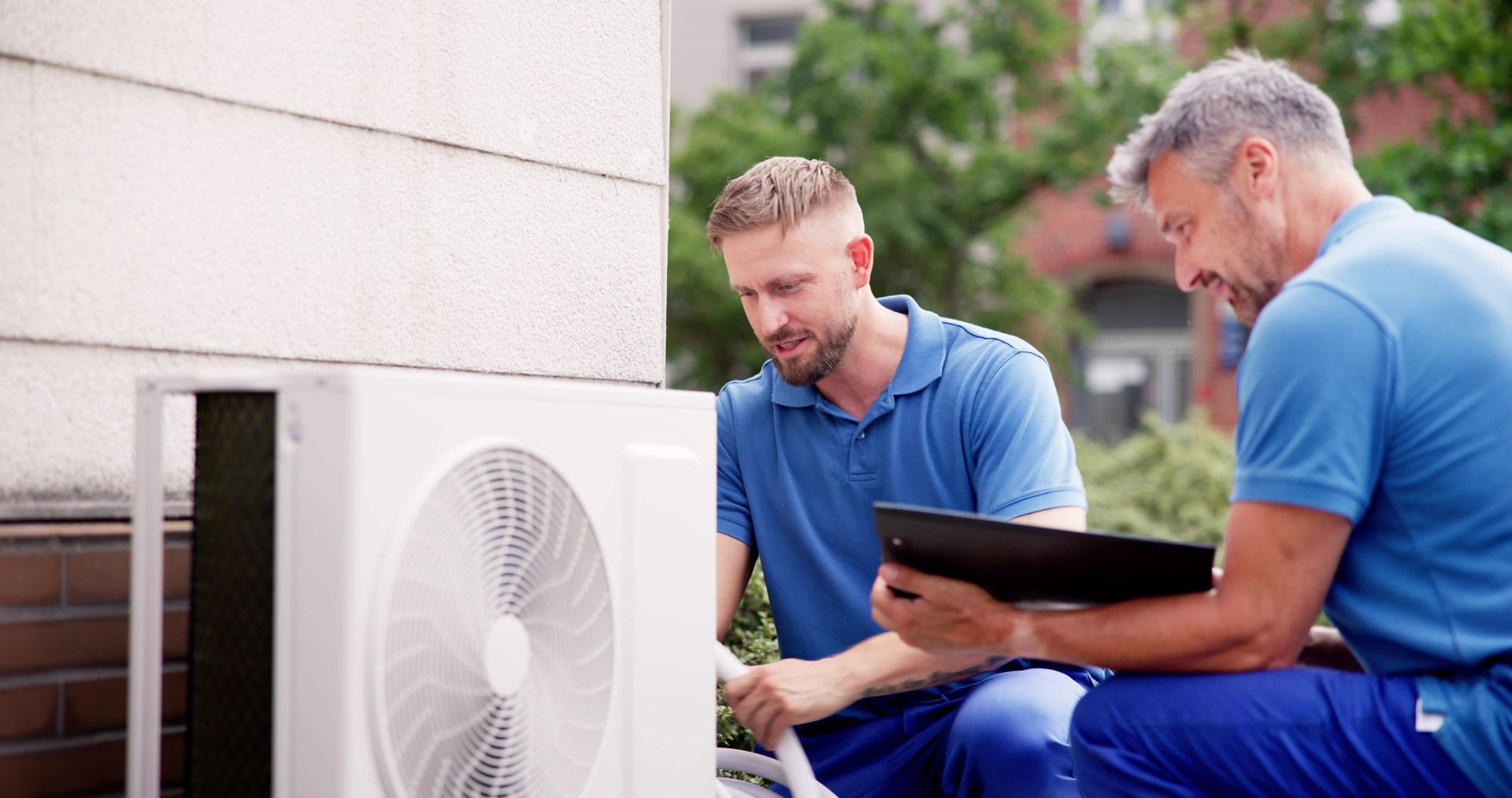 Two technicians in blue uniforms work together to inspect an outdoor HVAC unit, one checking the machine with a tablet.