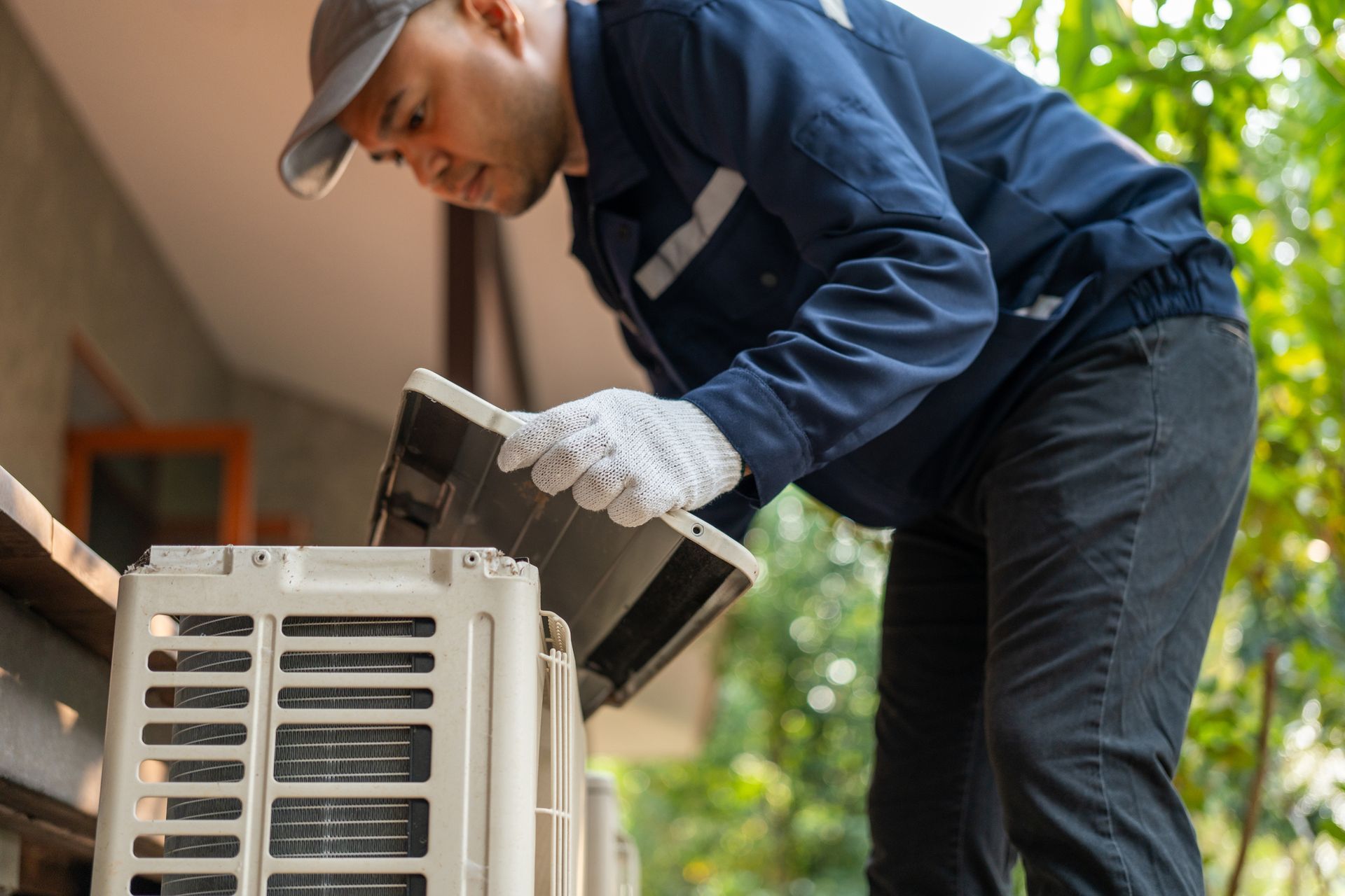 A worker wearing a cap, gloves, and blue uniform services an outdoor air conditioning unit.