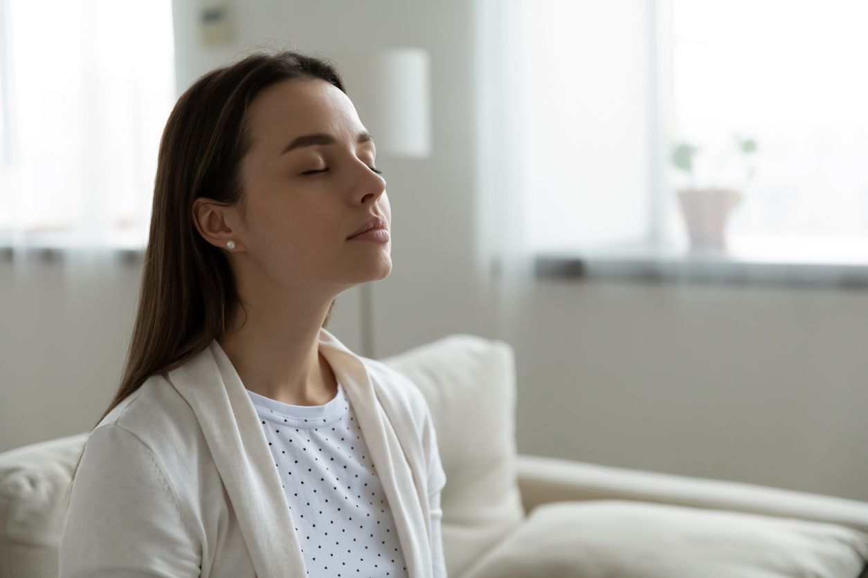 A person with eyes closed, appearing calm and relaxed, sitting on a couch in a bright, softly lit indoor space.