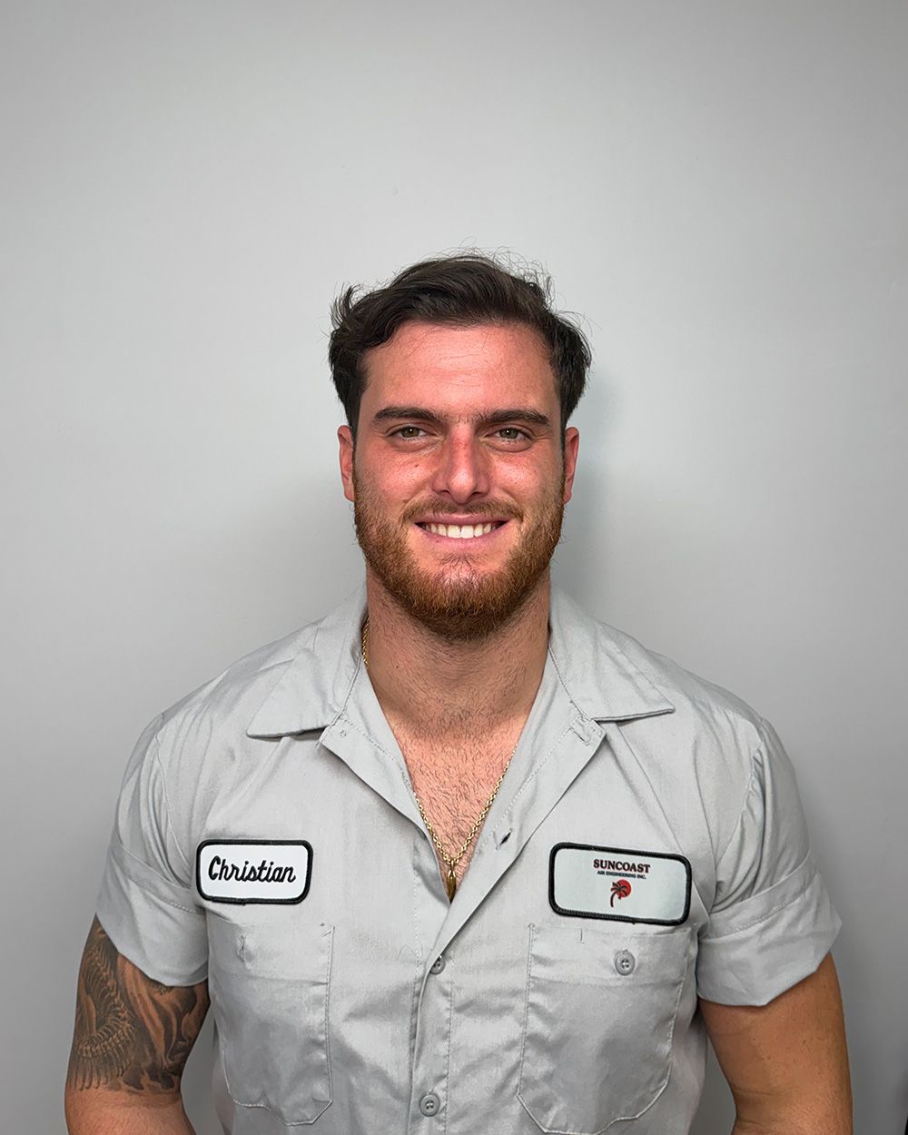 A person with a beard, wearing a short-sleeved light gray collared work shirt with patches, smiling against a gray wall.