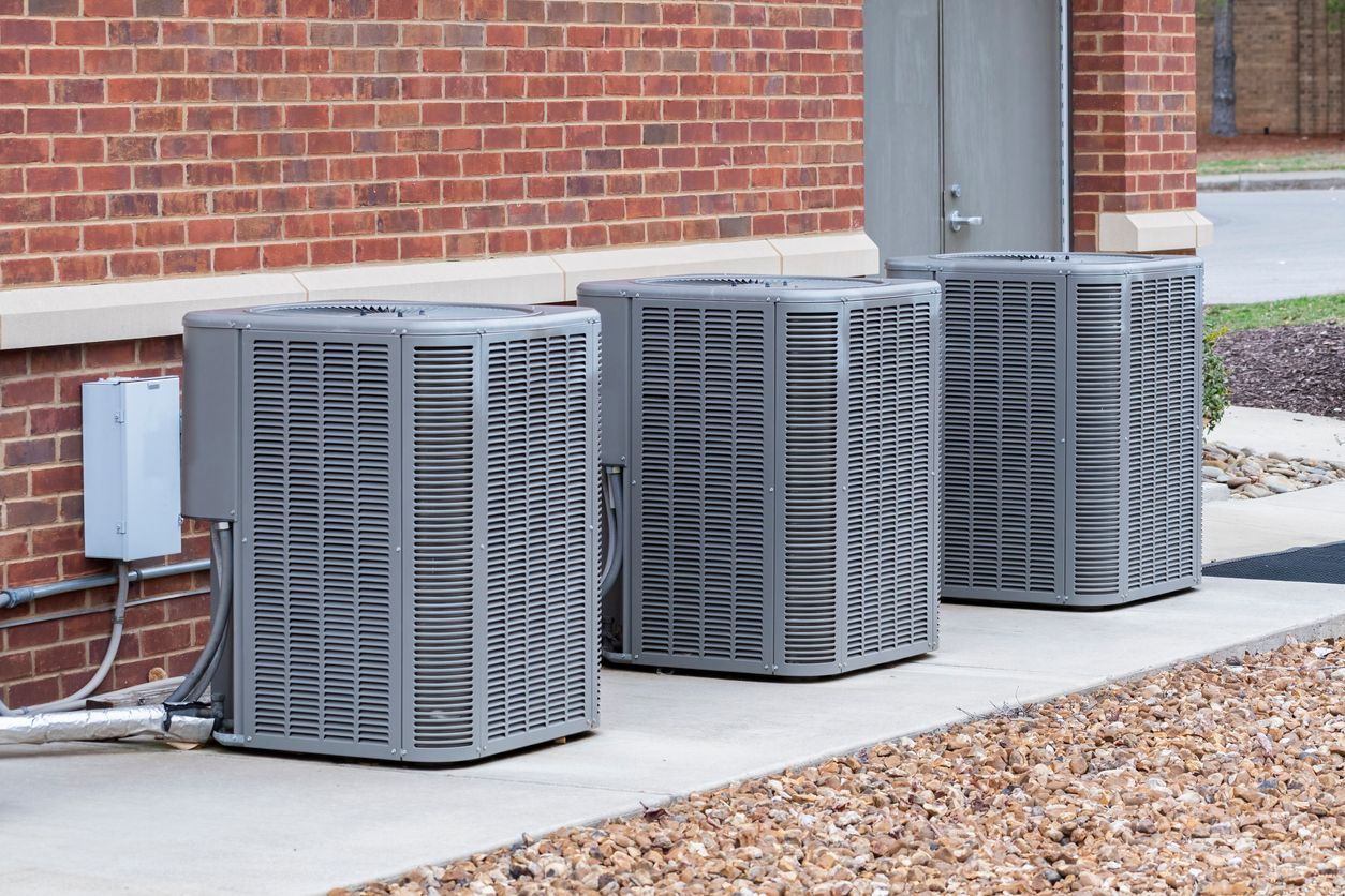 Three grey HVAC condenser units lined up on a concrete pad against a red brick building.