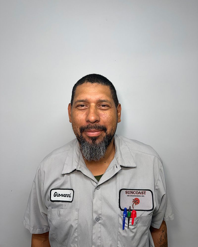 A person in a light grey work uniform with a beard and name patch, standing against a plain light grey background.