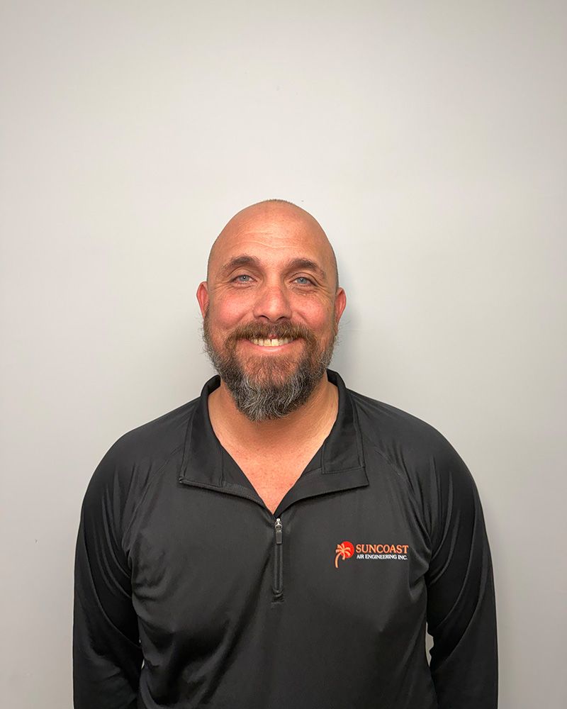 A smiling person with a beard, wearing a black long-sleeved athletic zip-up shirt against a plain light gray background.