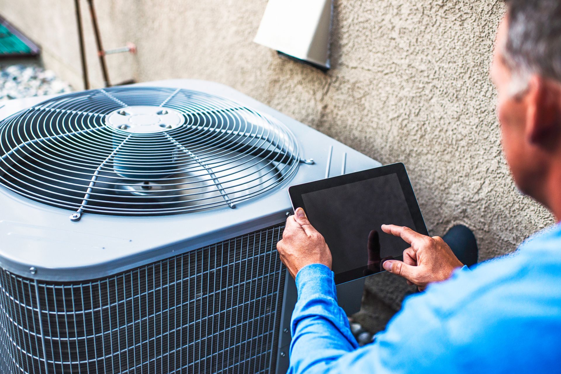 A technician in a blue shirt uses a tablet to inspect an outdoor air conditioning unit.