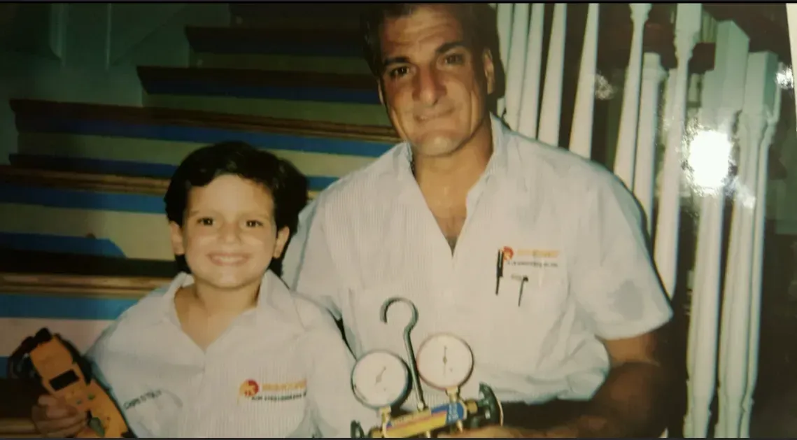 A child and an adult wearing matching work shirts pose on a staircase, holding HVAC diagnostic tools.