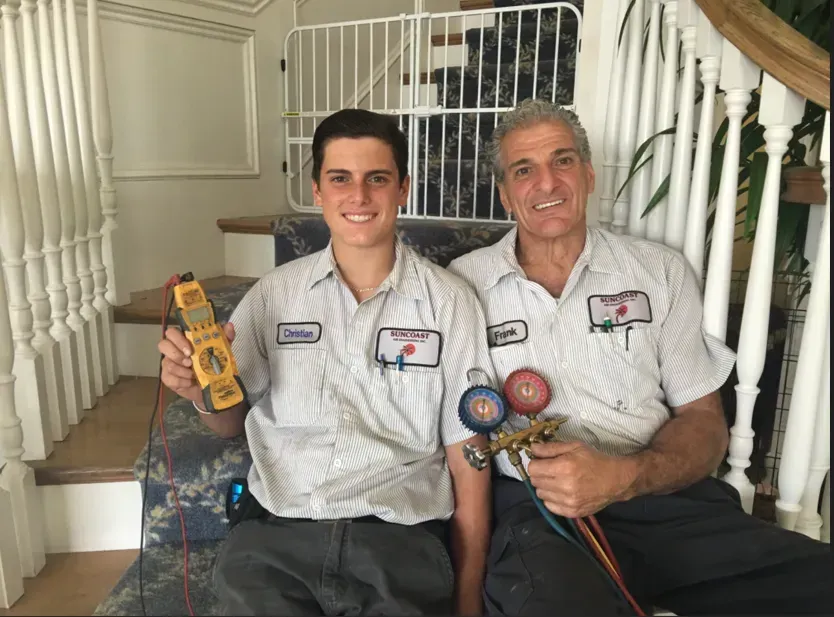 Two Suncoast Air Engineering technicians in work uniforms sit on stairs, one holding a multimeter.