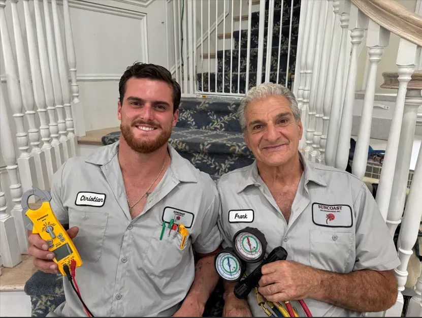 Two Suncoast Air Engineering technicians in gray work shirts hold tools on stairs, smiling at the camera.