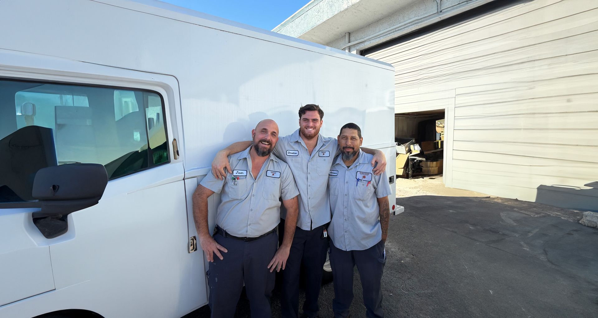 Three Suncoast Air Engineering technicians in matching grey uniforms stand in front of a white truck outside a warehouse.