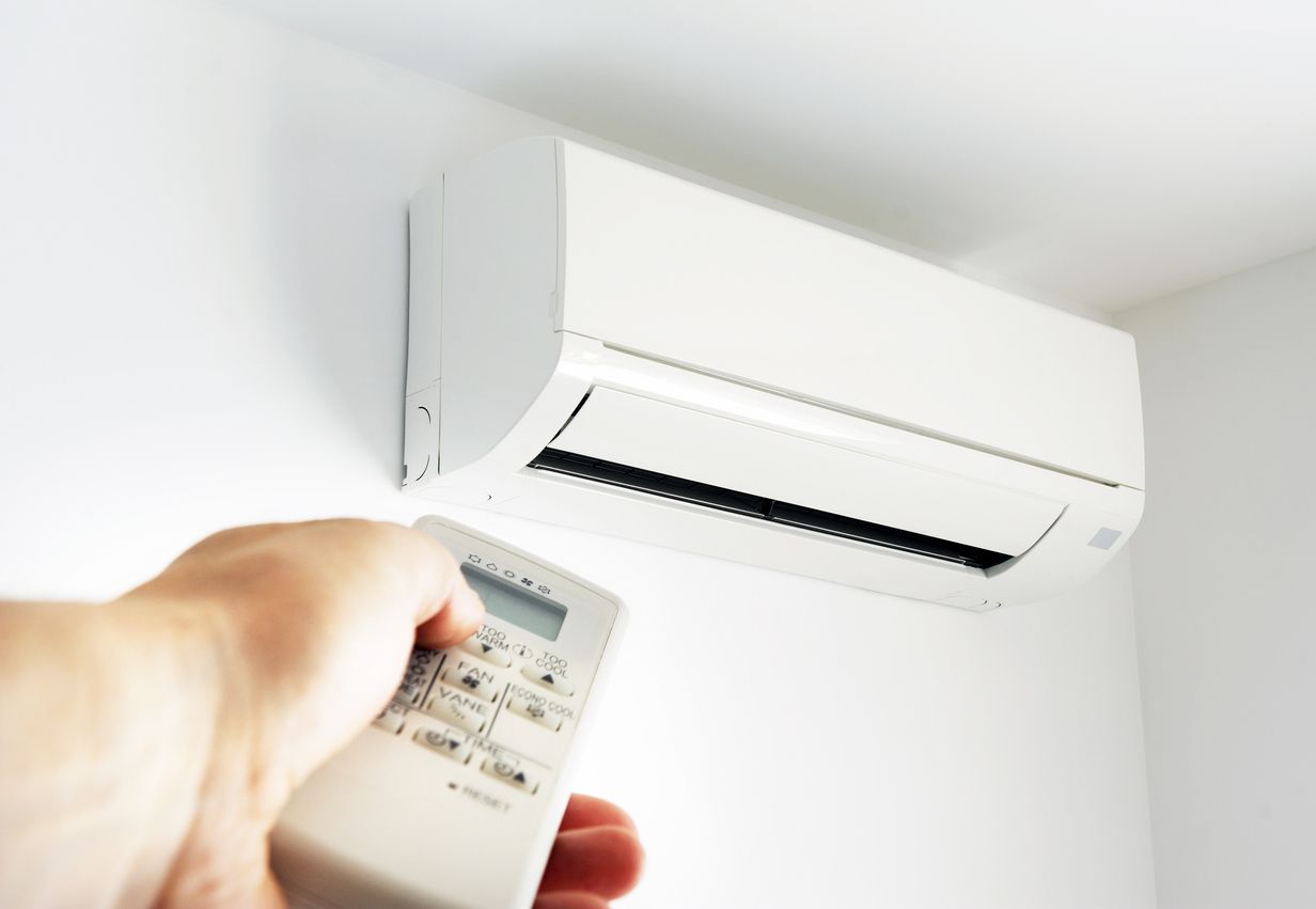 A person's hand holds a remote control pointed toward a white wall-mounted air conditioner unit in a room.