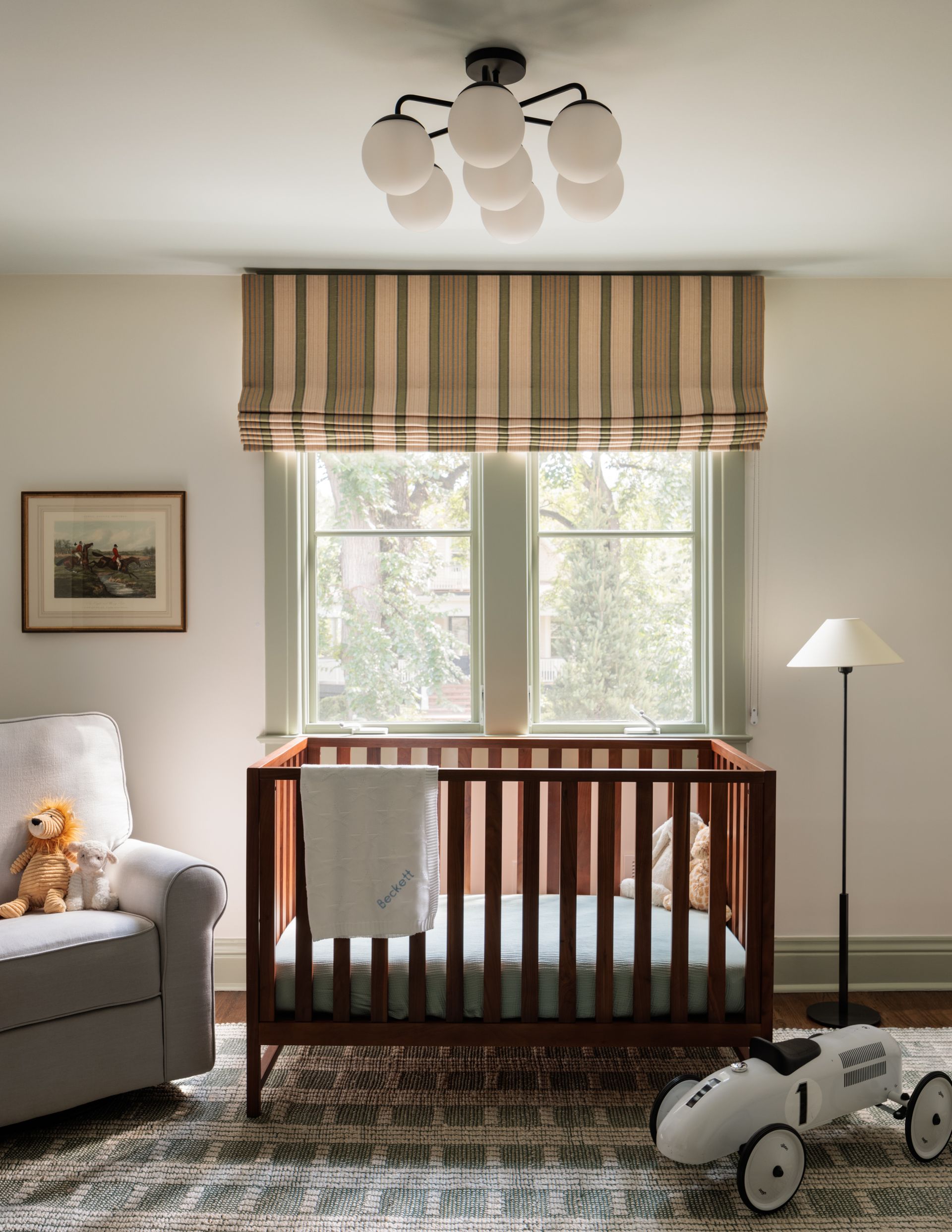 A nursery with a crib a chair and a toy car and roman shades with stripes