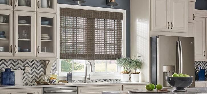A kitchen with white cabinets , a refrigerator , a sink , and a bowl of apples with woven wood blinds on the counter.