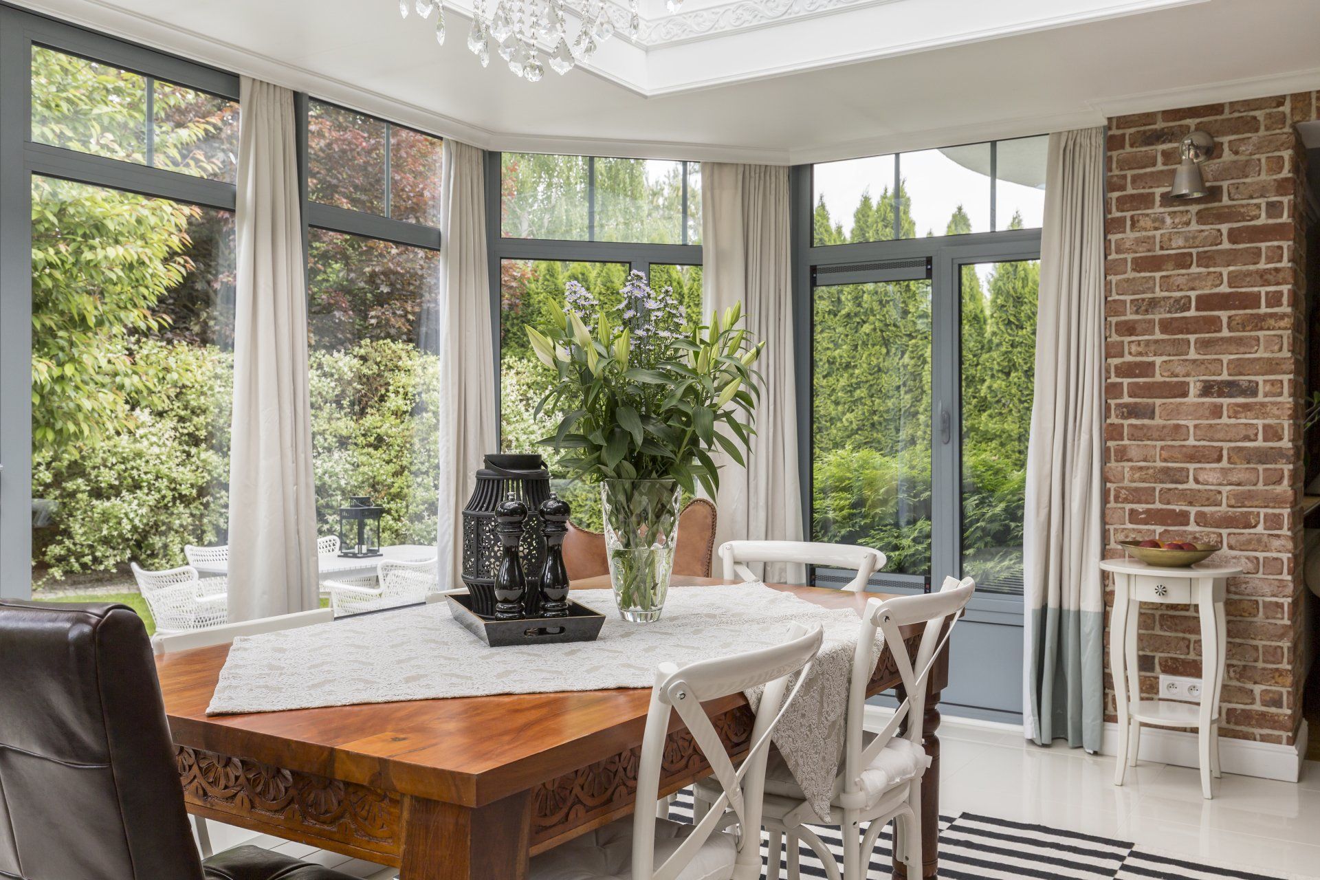 A dining room with a wooden table and chairs and a chandelier and white drapes in a screened porch