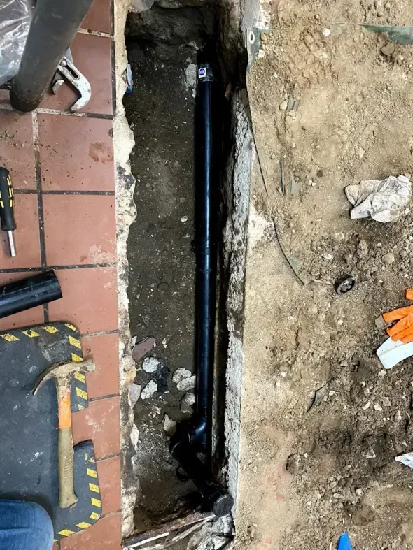 Plumbing work: black pipes in a trench next to red-tiled floor. Tools and dirt are present.